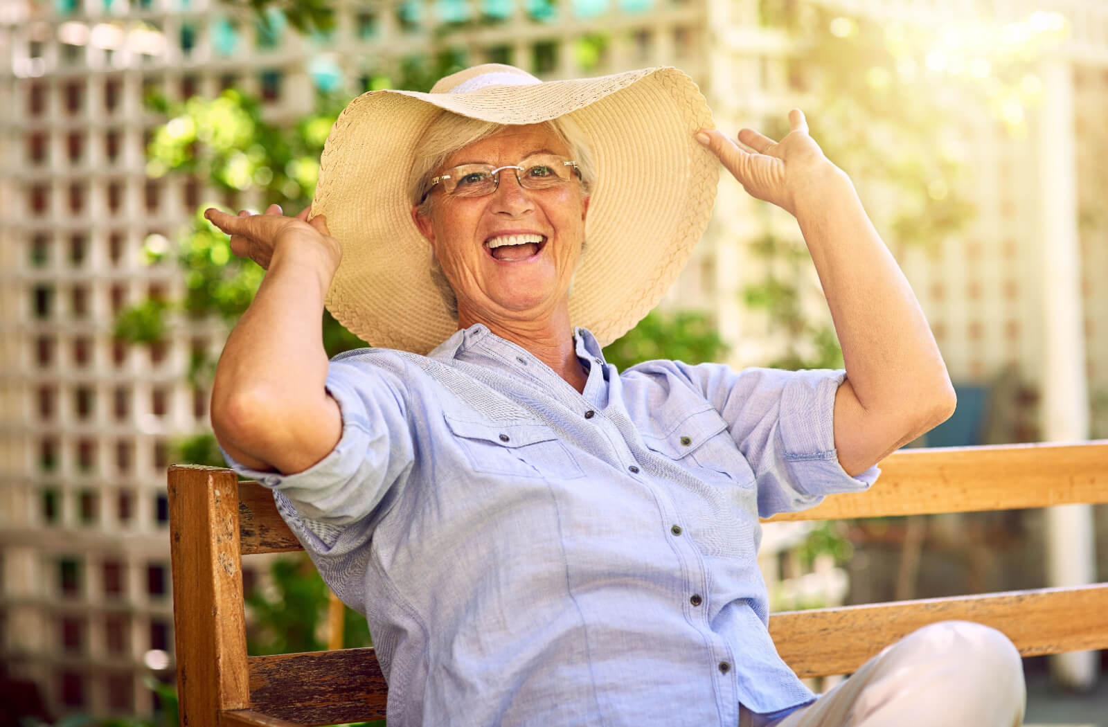An older adult in a sunhat raises their hands and laughs while sitting outdoors on a bench in a garden in senior living